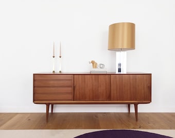 May include: A mid-century modern teak sideboard with three drawers and two doors. The sideboard has a gold lamp with a white shade, a small wooden elephant figurine, and two candlesticks on top.
