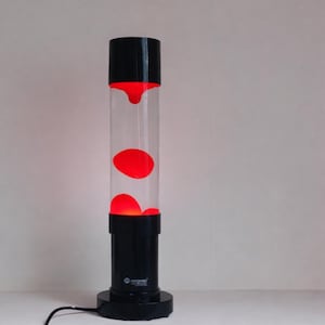 Könnte beinhalten: Eine schwarze und transparente Lavalampe mit roter Lava. Die Lampe hat ein zylindrisches Glasrohr mit rotem Wachs im Inneren und einen schwarzen Sockel und Deckel. Die Lampe ist eingesteckt und eingeschaltet, wobei die rote Lava beleuchtet wird.