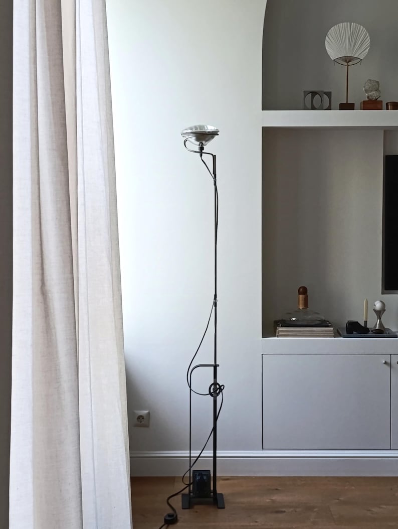 May include: A black floor lamp with a silver shade and a unique design. The lamp is in a room with white walls and a white curtain.