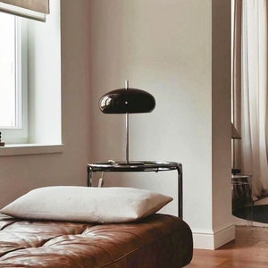 May include: A brown leather sofa with a white pillow and a black lamp on a glass side table. The sofa is in a room with a white wall and wooden floor.