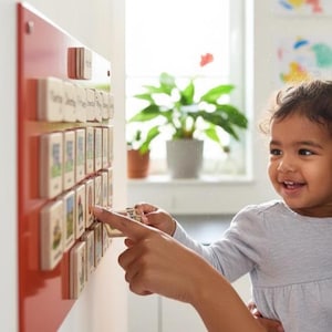 May include: A red magnetic board with wooden blocks featuring illustrations and text. A child points at a block while smiling. The board is mounted on a white wall, with a window and plants in the background.