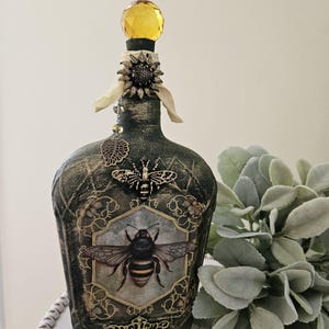 May include: Decorative bottle with a yellow crystal stopper, adorned with bee and sunflower embellishments. The bottle features a detailed bee illustration and ornate gold accents, creating a vintage aesthetic. The bottle is approximately 20 cm tall.
