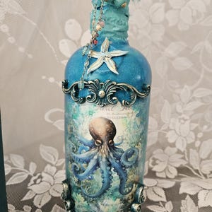 May include: A decorative glass bottle painted in shades of blue, featuring a central octopus illustration, a white starfish accent, and ornate metal filigree. The neck is wrapped in blue fabric with hanging beads and a clear crystal topper.