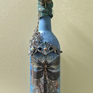 May include: A blue glass bottle decorated with a large dragonfly graphic, floral motifs, and ornate bronze-colored filigree accents. The neck is wrapped in green fabric with a dangling crystal chain, a metal dragonfly charm, and a faceted blue topper.
