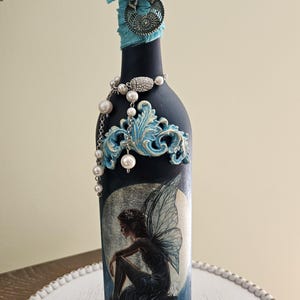May include: A matte black glass bottle decorated with a fairy illustration, a blue crystal topper, light blue fabric ribbon, a silver-toned metal charm, and a string of white faux pearls. Ornate light blue scrollwork accents the top and base.