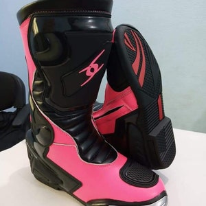 May include: Pink and black motorcycle boots with silver accents. The boots have a black leather upper with a pink leather lower section. The boots have a black zipper on the side and a black strap across the top.