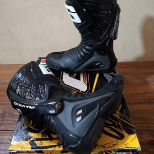 May include: A pair of black leather motorcycle boots with a gray and black design. The boots have a white logo on the side and the text "gaerne" on the ankle. The boots are sitting on a yellow and black box with the Gaerne logo.