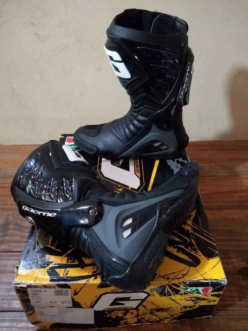 May include: A pair of black leather motorcycle boots with a gray and black design. The boots have a white logo on the side and the text "gaerne" on the ankle. The boots are sitting on a yellow and black box with the Gaerne logo.