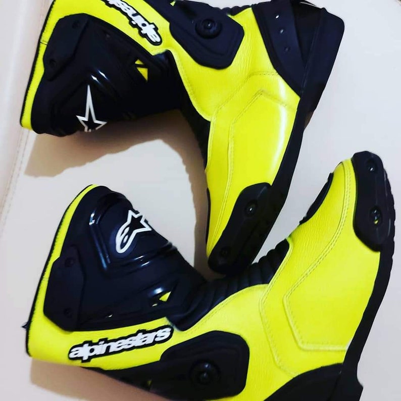 May include: A pair of yellow and black motorcycle boots with the Alpinestars logo. The boots have a black leather upper and a yellow leather lower. The boots have a black rubber sole and a black buckle.