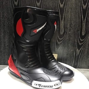 May include: A pair of black leather motorcycle boots with red accents and the Dainese logo. The boots have a silver toe cap and a black rubber sole.