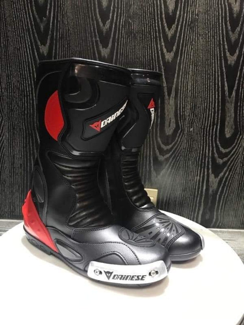 May include: A pair of black leather motorcycle boots with red accents and the Dainese logo. The boots have a silver toe cap and a black rubber sole.