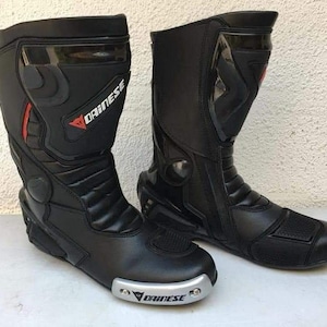 May include: A pair of black leather motorcycle boots with a silver Dainese logo on the side. The boots have a zipper on the side and a buckle on the top.