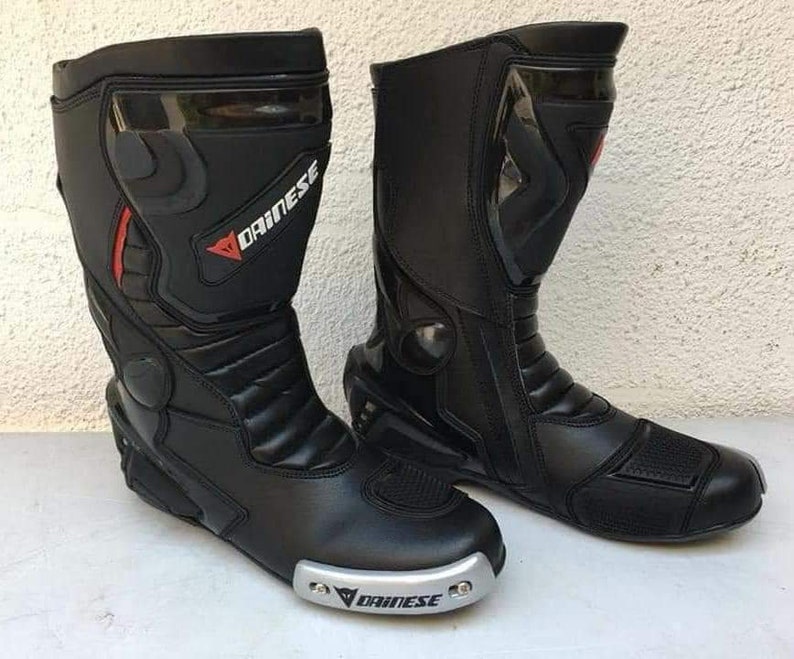 May include: A pair of black leather motorcycle boots with a silver Dainese logo on the side. The boots have a zipper on the side and a buckle on the top.