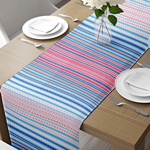 May include: A blue and pink striped table runner with a geometric pattern. The runner is on a wooden table with white plates and silverware.