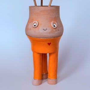 May include: A quirky terracotta vase with a smiling face, orange trousers, and three pom-pom flowers. The vase has a heart detail and is a unique decorative piece. The pom-poms are red, yellow, and pink.