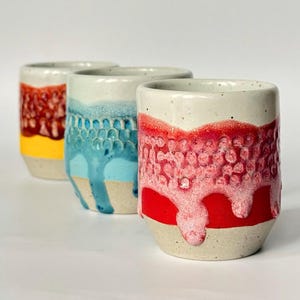 May include: Three ceramic tumblers with textured, dripped glaze in red, blue, and yellow. The tumblers have a speckled, off-white base and a cylindrical shape, perfect for beverages.