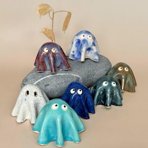 May include: A collection of seven small ceramic ghost figurines in various colours including turquoise, dark blue, white with crackle glaze, purple, and brown. Each ghost features two small black and white eyes and a draped, flowing shape.
