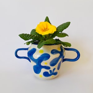 May include: A ceramic vase with two handles, decorated with blue abstract shapes and yellow dots. The vase holds a vibrant yellow flower and green foliage. The vase is on a white background, creating a colorful display.