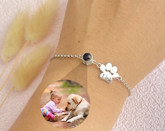 May include: Silver bracelet with a paw print charm engraved with the word "Link" and a black gemstone. The bracelet is on a person's wrist and there is a photo of a child and a dog in the background.