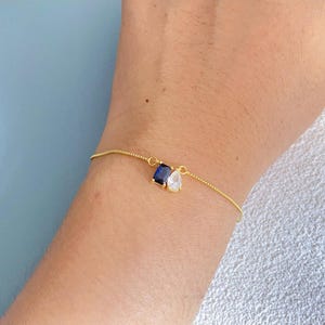 Könnte beinhalten: Ein zartes goldenes Armband mit zwei tropfenförmigen Edelsteinen: einem dunkelblauen Saphir und einem klaren Zirkonia. Die Edelsteine hängen an einer feinen Goldkette, wodurch ein minimalistisches Design entsteht. Das Armband wird am Handgelenk getragen.