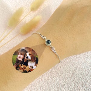 Op de afbeelding: Een zilveren armband met twee engelenvleugelcharms en een zwart fotomedaillon. Het medaillon bevat een foto van een kind en een hond.