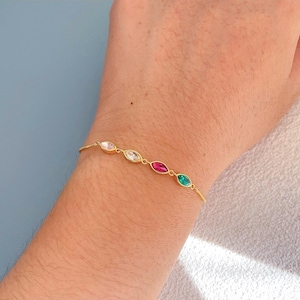 Könnte beinhalten: Ein goldenes Armband mit einer feinen Kette und fünf ovalen Edelsteinen in den Farben Klar, Weiß, Rot und Grün. Das Armband wird an einem Handgelenk gezeigt, um das Design und die Farben hervorzuheben.