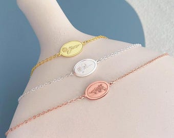 May include: Three delicate bracelets in gold, silver, and rose gold. Each features an oval pendant with a floral design. The bracelets are displayed on a white surface against a blue background.