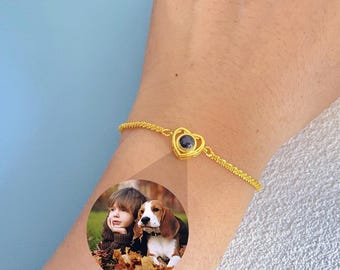 May include: Gold-colored bracelet with a heart-shaped pendant. The pendant has a dark center and is connected to a delicate chain. A circular image of a child and a dog is visible.