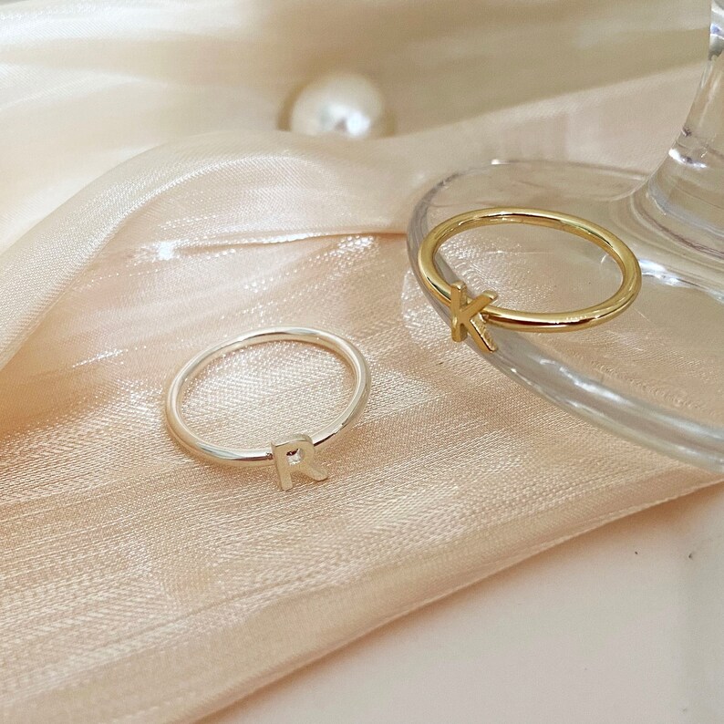 Peut inclure: Deux bagues simples en or et en argent avec les lettres initiales 'K' et 'R' sur un fond de tissu blanc.
