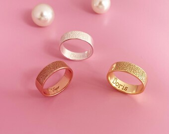 May include: Three personalized rings with textured surfaces. The rings are silver, gold, and rose gold. The names "Oscar", "Doris", and "Viger" are engraved on the inside of the rings.