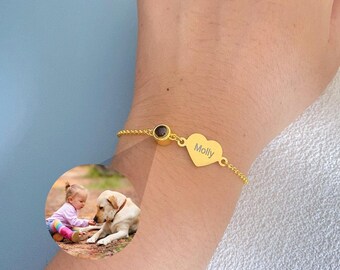 Puede incluir: Una pulsera dorada con un dije en forma de corazón grabado con el nombre "Molly". La pulsera también presenta una pequeña piedra redonda de color oscuro y una delicada cadena. Una pequeña foto de un niño y un perro está en el fondo.