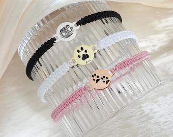 May include: Three bracelets with paw print charms. The bracelets are black, white, and pink. The charms are silver, gold, and rose gold. The gold charm has the word "Moi" engraved on it.