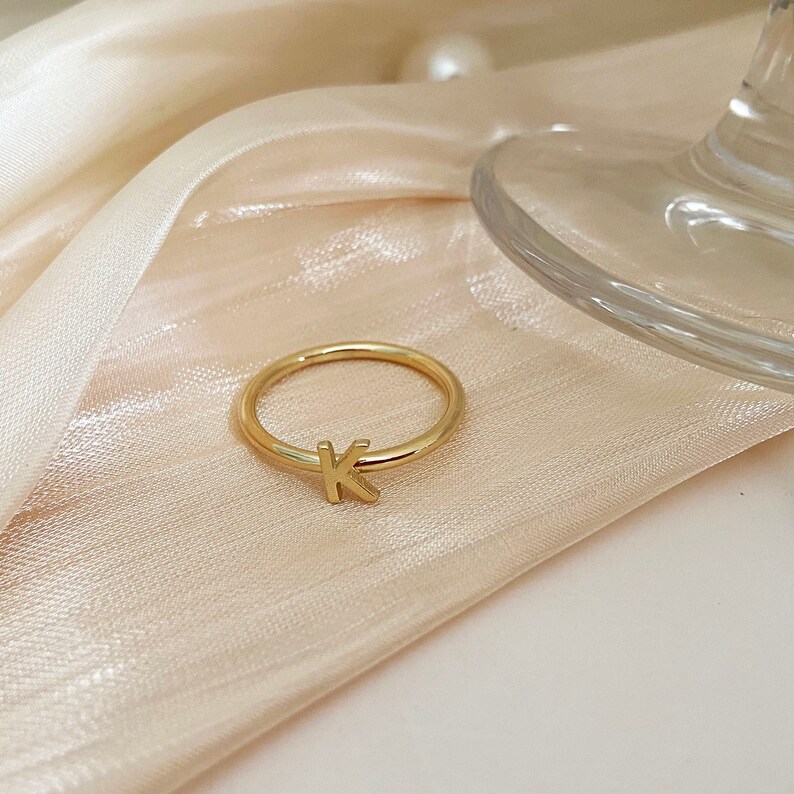 Peut inclure: Une bague en or avec la lettre "K" dessus. La bague est sur un fond de tissu beige clair.