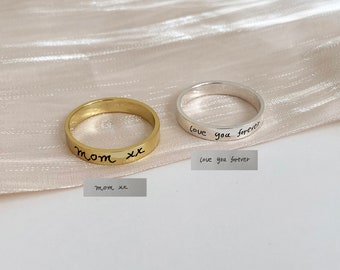 Puede incluir: Dos anillos finos de oro y plata con mensajes escritos a mano. El anillo de oro dice "mom xx" y el anillo de plata dice "love you forever".
