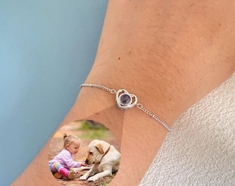 May include: A silver bracelet featuring a heart-shaped pendant with a small, dark-colored stone. The bracelet is on a wrist, with a circular image of a child and a dog. The bracelet's chain is delicate and the heart pendant is the focal point.