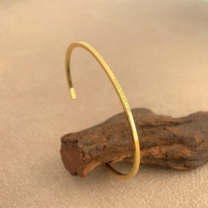 Puede incluir: Una pulsera de brazalete abierta de color dorado. La pulsera descansa sobre un trozo de madera. El texto "Believe that good is love" está grabado en la pulsera. La pulsera tiene un diseño simple y elegante.