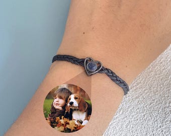 Puede incluir: Un brazalete trenzado negro con un dije en forma de corazón con una piedra azul. El dije está en la muñeca de una mujer. Una foto de un niño y un perro es visible en el brazo de la mujer.