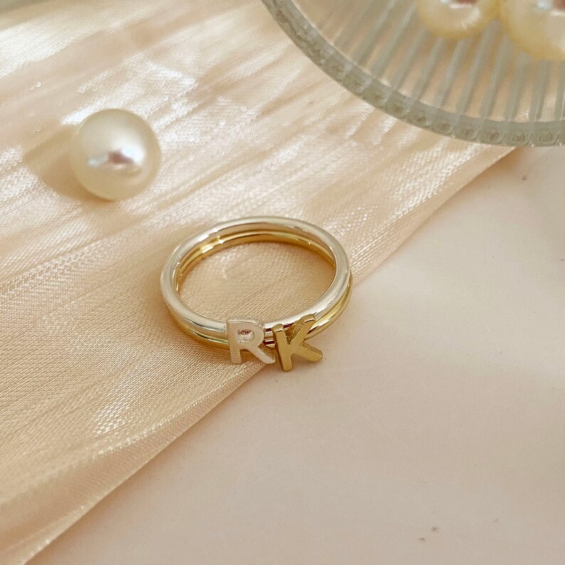 Peut inclure: Une bague en or et argent avec les initiales "RK" dessus. La bague est sur un fond de tissu beige clair.