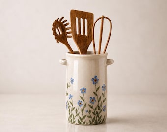 Op de afbeelding: Een cilindervormige keramische keukengereihouder met twee kleine handvatten aan de zijkant, voorzien van een handgeschilderd patroon van blauwe bloemen en groene stengels. In de houder staan drie houten keukengerei: een pastavork, een spatel met sleuven en een garde.