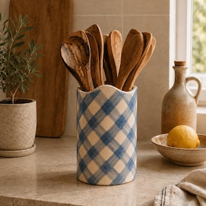 May include: A cylindrical ceramic utensil holder featuring a blue and white gingham check pattern, filled with several wooden spoons. The holder has a scalloped top edge and is positioned upright, displaying a hand-painted style finish.
