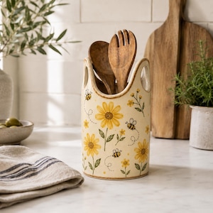 May include: A cream-coloured ceramic utensil holder featuring a painted pattern of yellow sunflowers, green stems, and small flying bees. The cylindrical container has two side cutouts for handles and holds two wooden kitchen spoons.