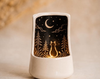 Op de afbeelding: Een crèmekleurige keramische kaarsenhouder met een gebogen achterpaneel en een zwarte binnenkant. Het ontwerp toont een gouden silhouet van een kat die in een bos onder een maansikkel en sterren zit, met een warm licht vanuit de basis.