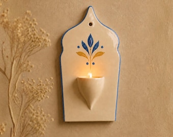 Op de afbeelding: Een crèmekleurige keramische wandkandelaar in de vorm van een spitsboog, voorzien van een blauw en geel bloemmotief en een klein bakje voor een waxinelichtje. De kandelaar heeft een gat aan de bovenzijde en een brandend kaarsje in het bakje.