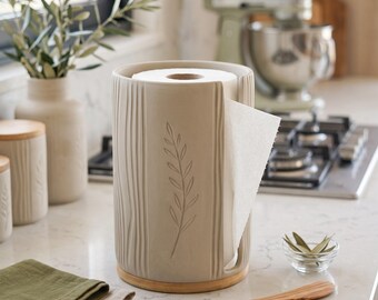 Op de afbeelding: Een cilindrische beige keramische keukenrolhouder met verticale ribbeltexturen en een uitgesneden bladmotief aan de voorzijde. De houder is voorzien van een ronde lichthouten voet en een verticale gleuf voor het afrollen van papier.
