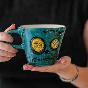May include: A teal ceramic mug featuring a hand-painted graphic of a face with two large yellow spiral eyes, a small triangular nose, and black stitched lines across the mouth and forehead. The mug has a wide handle and a smooth, glossy finish.