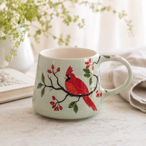 May include: A light green ceramic mug featuring a hand-painted illustration of a bright red cardinal bird perched on a dark brown branch with green leaves and small red berries. The mug has a wide, rounded body and a curved handle on the right side.