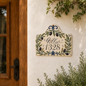 May include: A cream-colored ceramic house sign with a decorative arched top, featuring hand-painted green olive branches, dark purple olives, and blue scrollwork accents. The text Villa 1328 is written in black script and serif font.