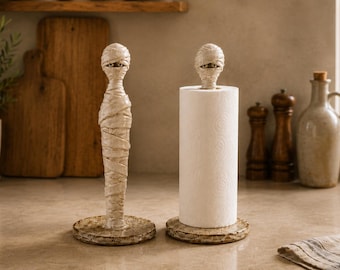 Op de afbeelding: Twee verticale keukenrolhouders met een beige mummiefiguur met omwikkelde bandages en zichtbare ogen aan de bovenkant. Elke houder heeft een ronde, getextureerde voet. Eén houder is leeg, de andere bevat een rol wit keukenpapier.