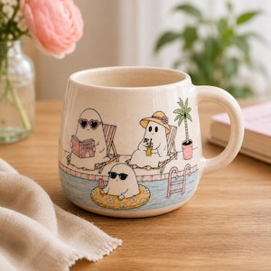 May include: A cream-colored ceramic mug featuring a colorful illustration of three ghosts relaxing by a swimming pool. One ghost wears heart-shaped sunglasses and reads a book titled Ghost Stories, another wears a sun hat, and one floats in a tube.