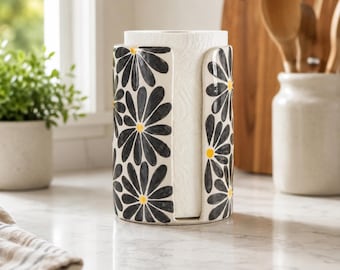 Op de afbeelding: Een cilindrische papieren handdoekhouder van keramiek met een witte basis en een herhalend patroon van zwarte madeliefachtige bloemen met gele harten. De houder heeft een verticale opening aan de voorzijde voor toegang tot de keukenrol.